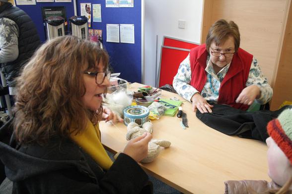 Mim, a woman with light skin tone and long curly brown hair, sewing up a small child's teddy which is now fully stuffed, while chatting to a child in a woolly hat. Across the table from her Ann, a woman with light skin tone and short brown hair, is sewing a black jacket and listening to the conversation.