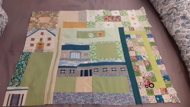 A large square of patchwork, depicting three stylised houses, a small store room and a chick coop, with areas of green grass and floral fabric flower beds. Fields are represented around two sides by pieced paper squares. In one field, a line of plain green squares among floral patterns is the trail left by a tractor, a motif which has been appliqued. 