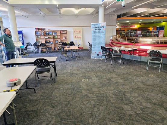 The entrance to Barry library with lots of tables and chairs laid out around it and a large sign for Repair Cafe Barry. At the door stands a man with light skin tone and short black hair with his hands on his hips surveying the scene.