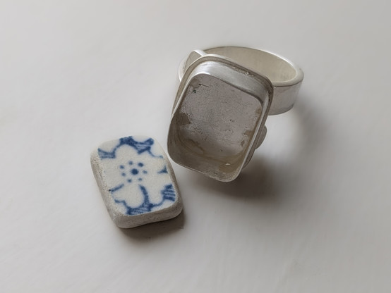 Colour photograph of a silver bezel ring setting on a hammered silver band. Next to it is a piece of floral blue sea pottery, which will eventually be set in the ring.