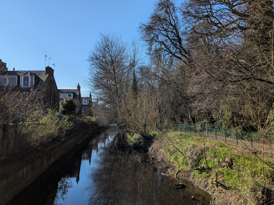 Colour photograph taken standing on a bridge across a stretch of water. To the left terraced stone houses, to the right green painted railings, a path, and trees. The water is still and reflects the houses and trees, and the sky is blue.
