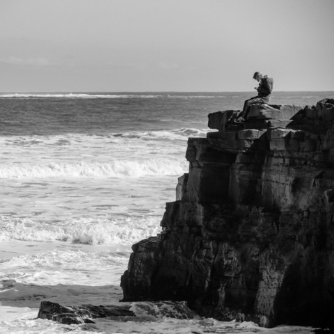 A black and white photograph. A seascape with a rough sea and the sky behind. On the right is a rocky outcrop with a figure sitting on top.