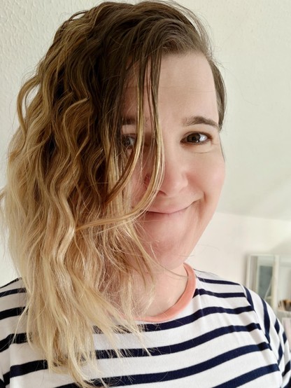 A picture of me smiling at the camera. I'm wearing a white top with black stripes. My hair is curly and blonde and partially covering my face.