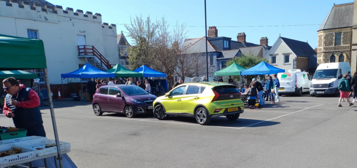 An image of Roath Market, Mackintosh Sports Club's car park in Keppoch Street. The crenulated white building to the rear left is that of the club. The photo was taken from the entrance and shows some of the stalls in the distance and one to the left, all under blue and green awings. There are many more stalls lined up along all four sides of the space but are out of the camera's view. Better composition next time!