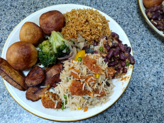A family-style West African meal from Mami Maggie's Recipes sits on a pale and brown-edged plate. The meal deal is shared, so an identical plated meal peeps out to the upper right. On the plate, clockwise from the top: Jollof rice, red beans, fried vegetable rice topped with hot sauce, a dollop of very hot sauce (this is Mme's plate, M NotLeafy had the rest), fried plantain and three puff puffs. A serving of mixed vegetables, including broccoli, red onions, peppers and cauliflower is in the centre of the plate. Both plates lie on a grey and brown speckled kitchen worktop.