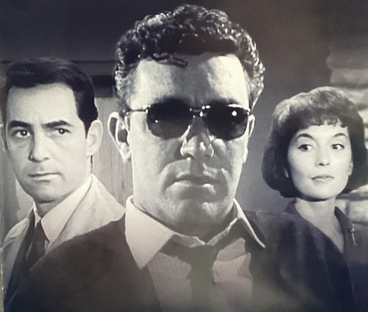 A man in dark glasses flanked by another man and a woman 