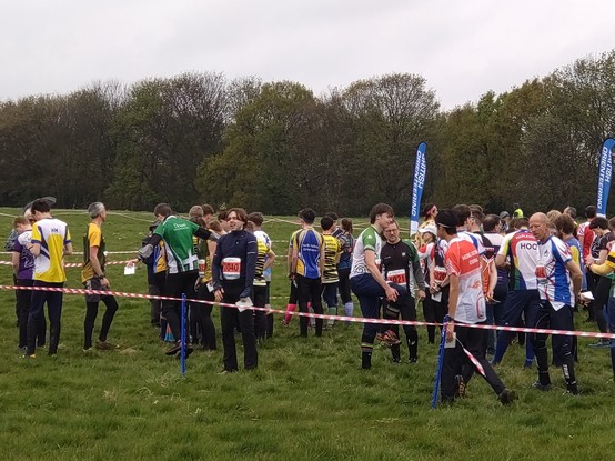Call up pre start for an orienteering relay - many colourful people in a wet field, forest in background