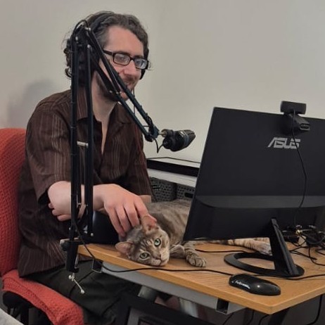 Chris from behind a desk, lightly stroking a cat, which is lying down where his keyboard should be.