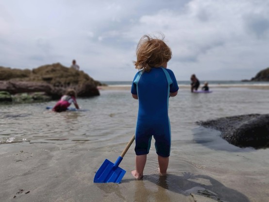 Picture of a toddler in a wetsuit holding a spade, and the background out of focus is a lovely seaside scene with pools and rocks.