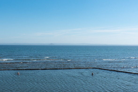 Colour photograph of a stretch of blue sea. A section at the front is surrounded by a stone wall, forming an outdoor swimming pool. The water in this area is still, and the heads of two swimmers are visible. The sea beyond has small white crested waves. The sky is blue.