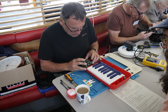 Kevin, a man with light skin tone and short black hair wearing glasses, sits at a table looking down at a circuit board which he is holding. On the table is a small red keyboard.