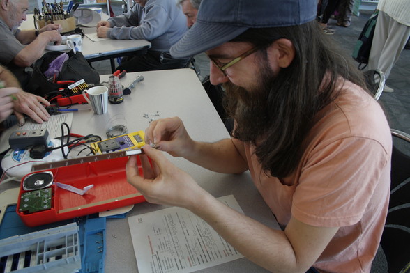 A person with light skin tone and long dark hair with a beard sits at a table. In their hands is a small circuit board which they are looking at intently.