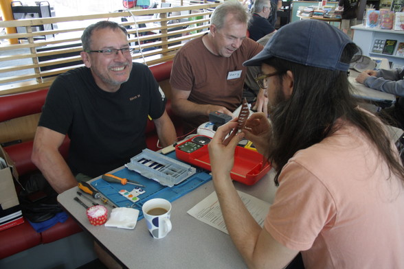 Kevin, a man with light skin tone and short black hair wearing glasses, sits at a table grinning broadly as the person across from him holds and looks at a circuit board.