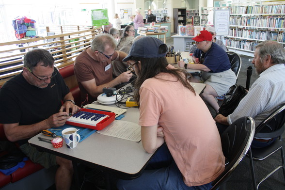 A table at Barry Repair Cafe with four people sat around it. In the foreground a small red keyboard is being worked on. In the background is another table where two people are waiting while their items are fixed. Rows of library books are dotted around.