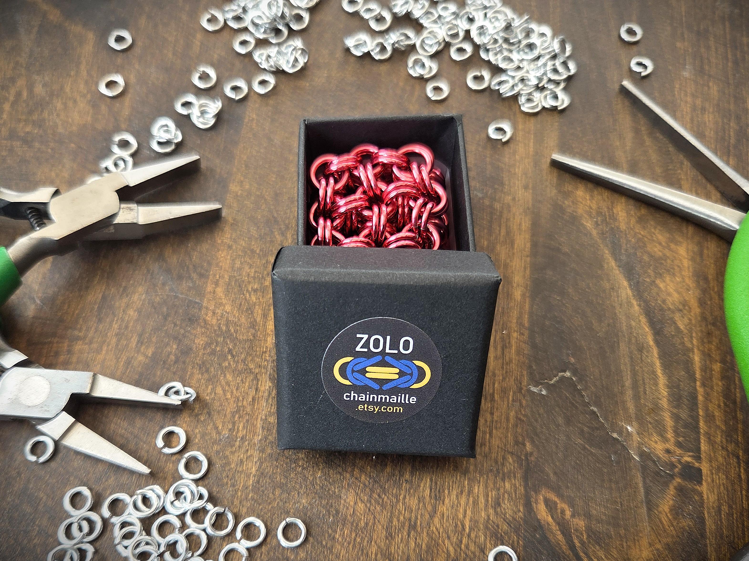A pink chainmaille fidget cube in a black gift box, on a dark wooden table, surrounded by silver jump rings and green-handled pliers