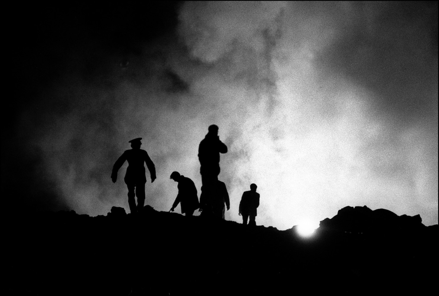 Exhausted miners and helpers are silhouetted against the arc lights and steam coming off the coal mine slurry after digging all day for survivors