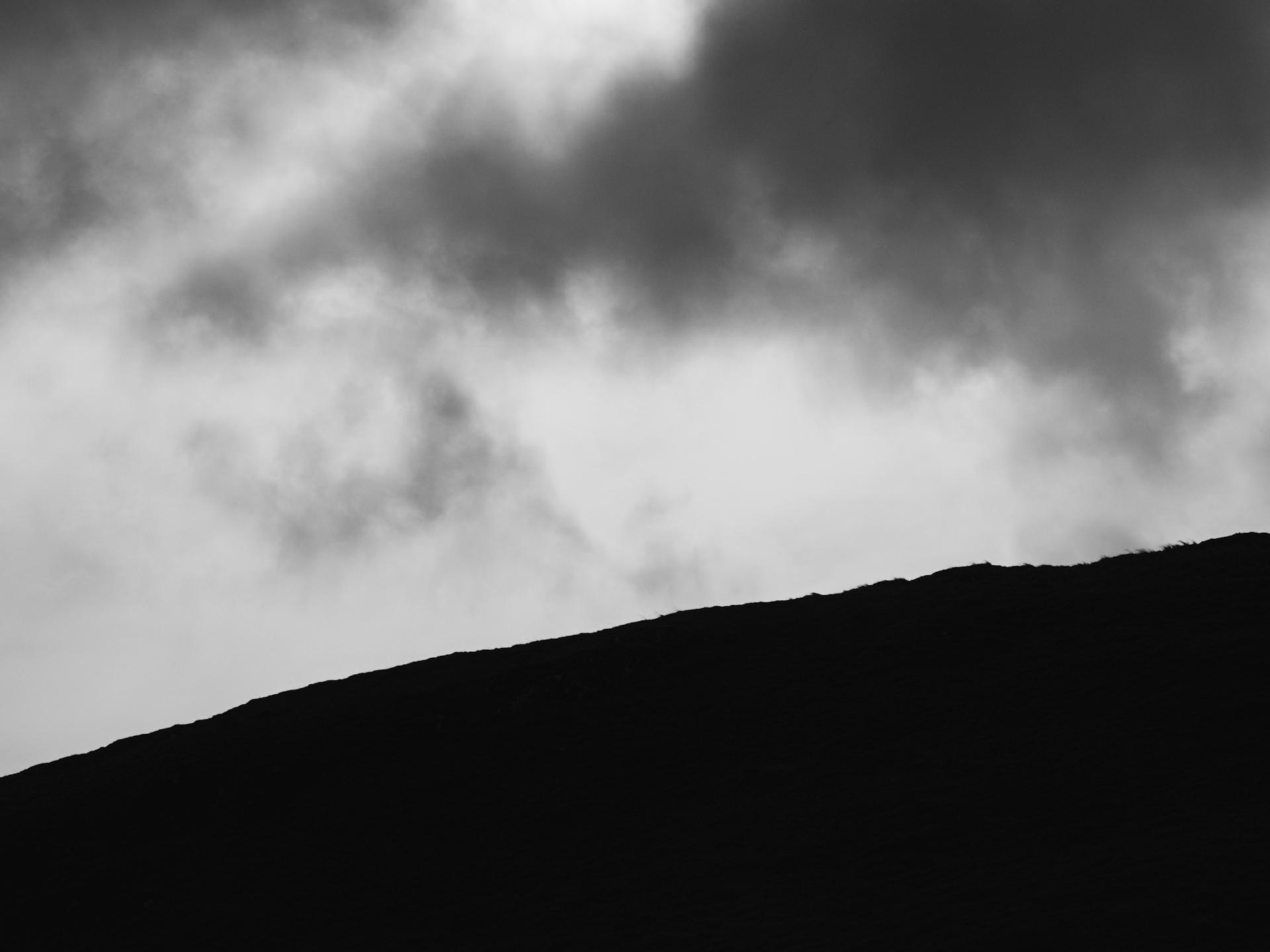 A silhouette of a hillside against a cloudy sky, presented in black and white.