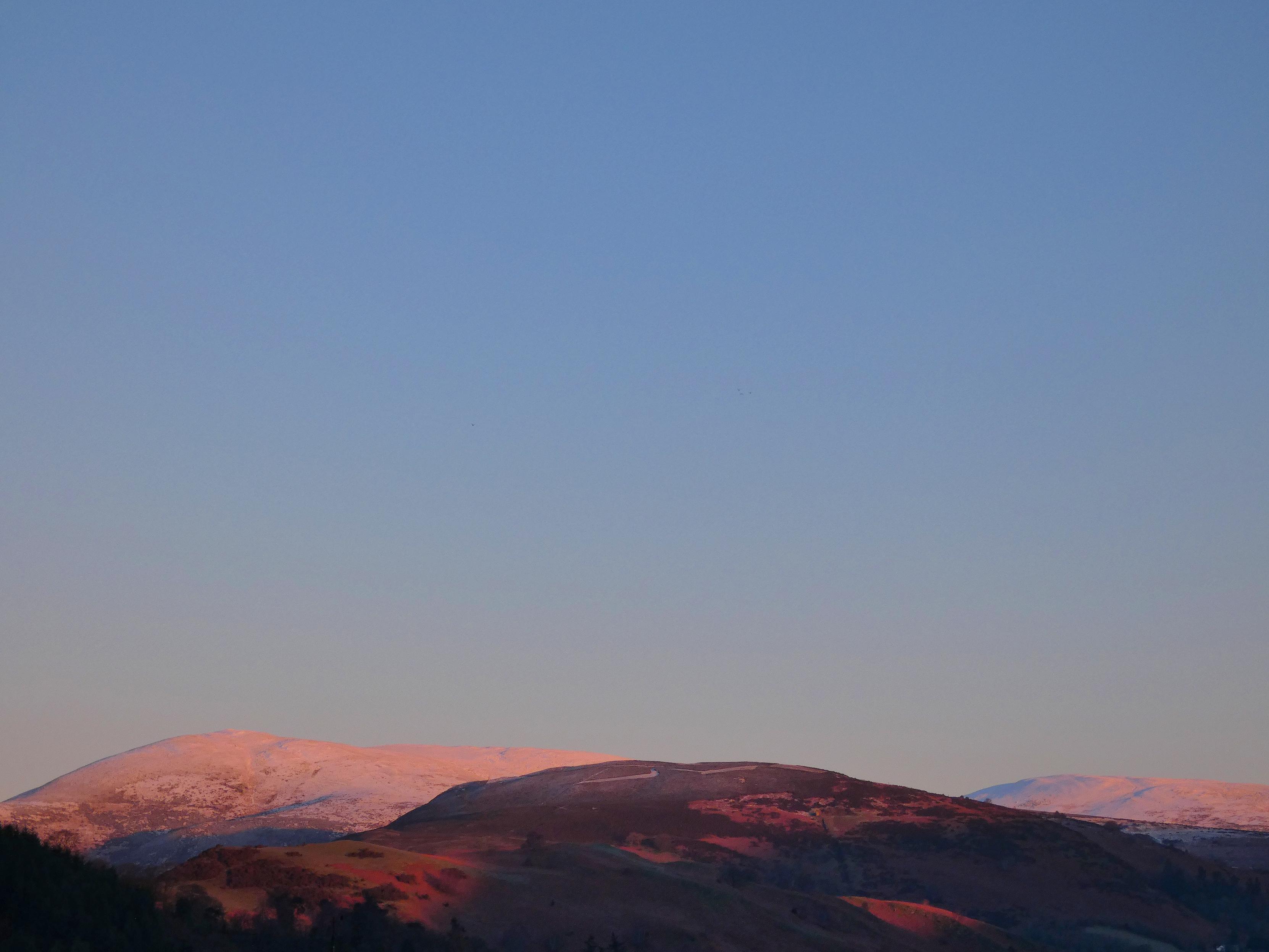 Snow capped distant hills lit in a rich golden light that enhances rust browns and oranges of their heathers, beneath wide clear blue skies