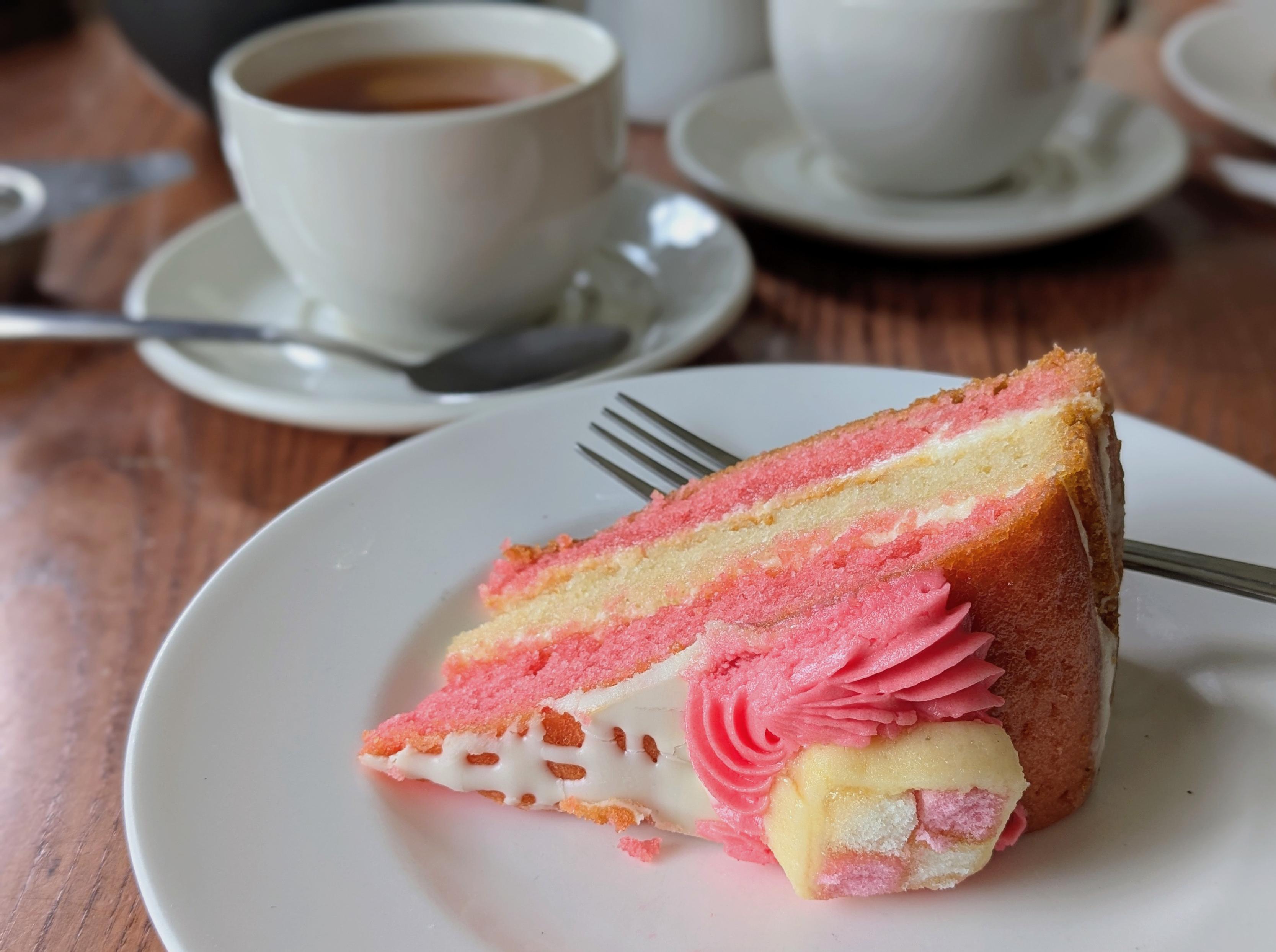 Colour photo of a big slice of pink and yellow layered sponge cake and a cup of black tea with white crockery.