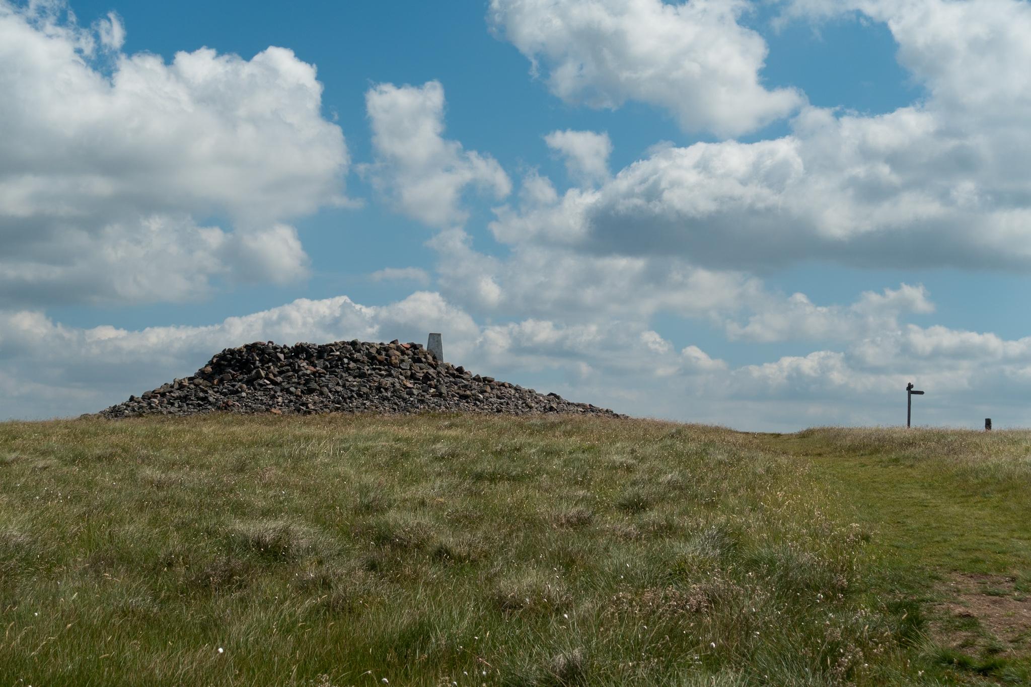 Colour photo of a large pile of stones, with a white trig point rising out of it. To the right is a fingerpost. There is a stretch of grass in the foreground. The sky is blue and full of puffy white clouds.