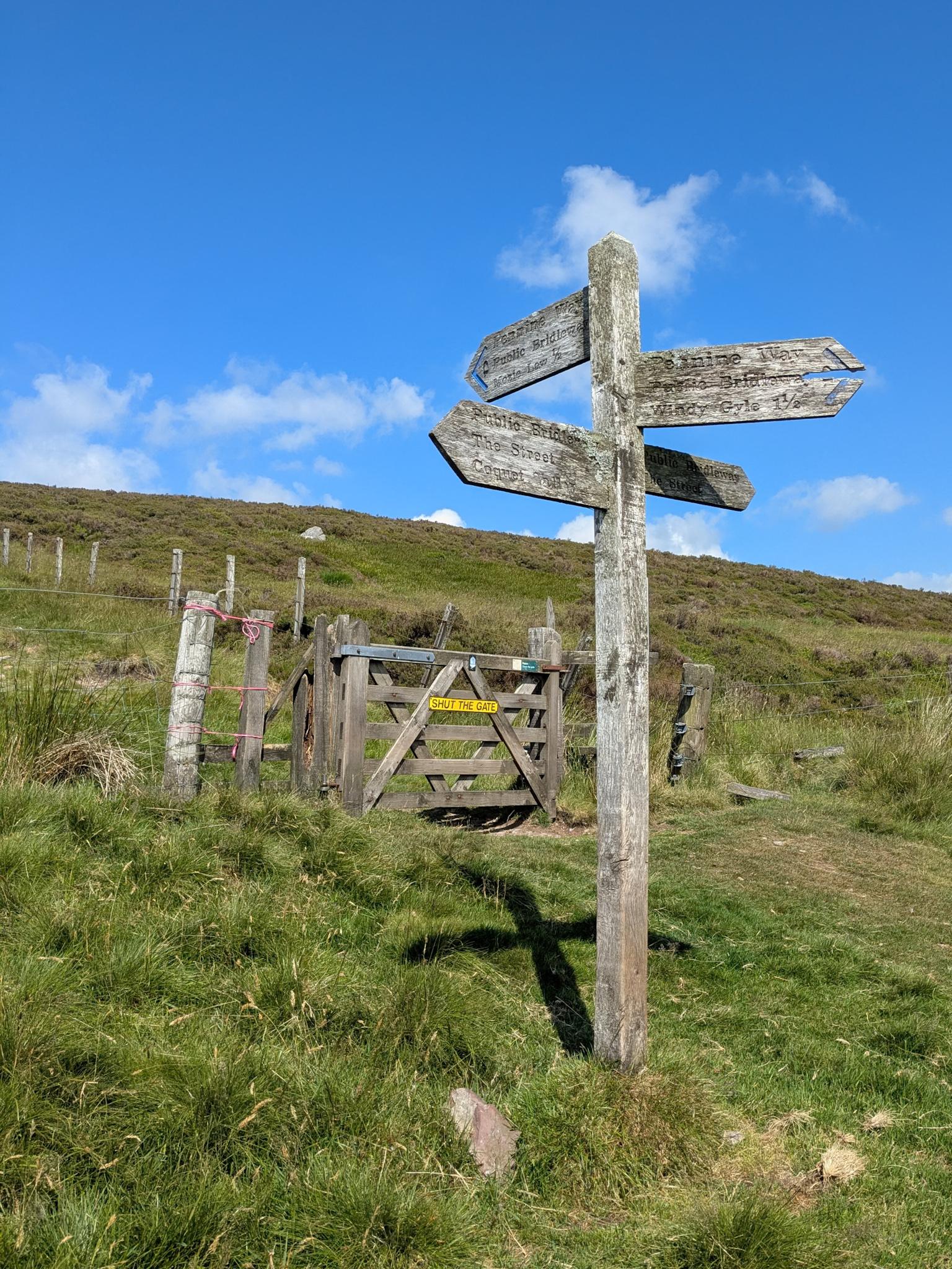 Colour phot of a wooden fingerpost pointing in four directions. It is set on a stretch of grass, with a fence and wooden gate behind. The sky is very blue.