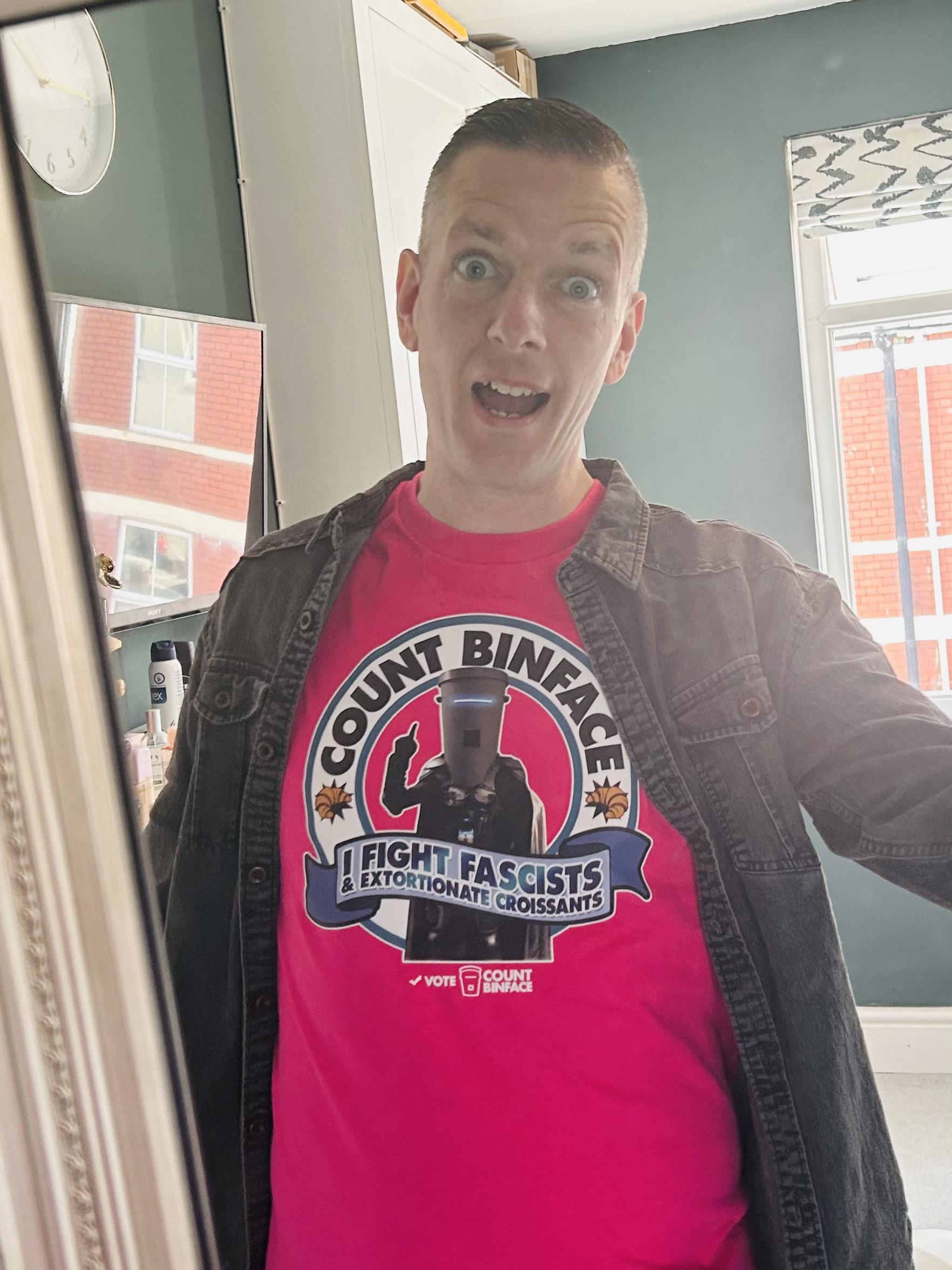 Ugly bloke with yellowing teeth and a gormless expression wearing a bright pink Count Binface t-shirt.