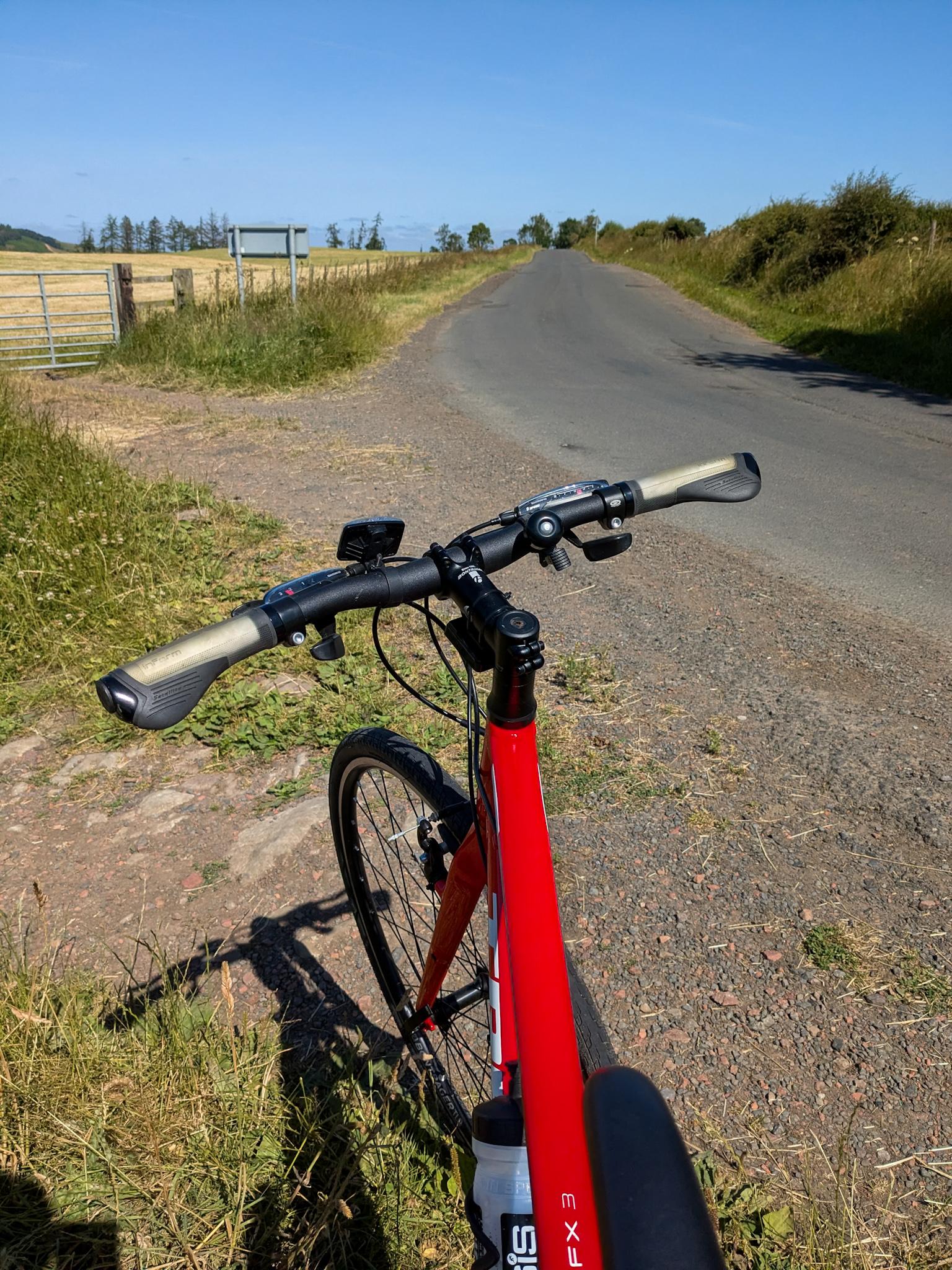 Colour photograph of the red crossbar, handlebars, and front wheel of a bike, pointing down a tarmac road with grassy verges. Blue sky.