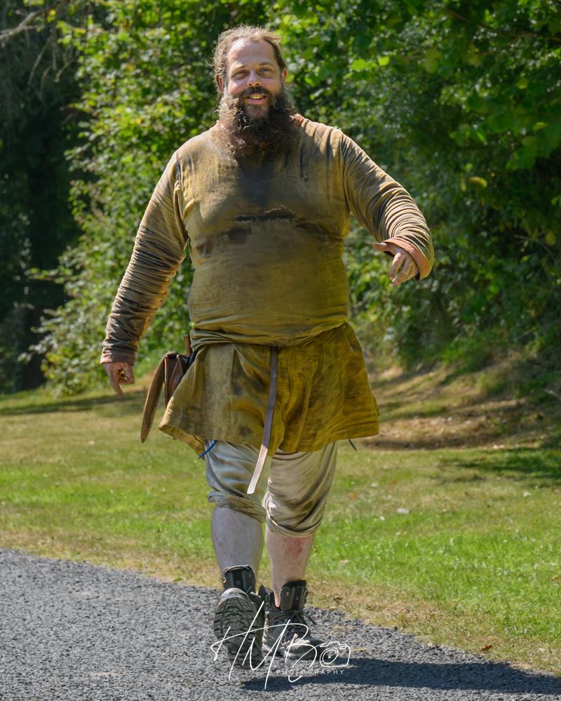 A giant of a bearded, smiling man in medieval style tunic, knee length trews, strides along a gravel path. He has a leather belt on his tunic.