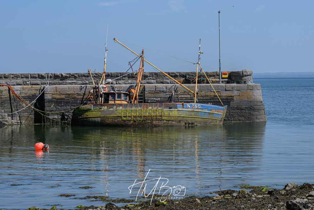 A boat that has seen better days, in a small harbour. There is a lot of greenish algae on the hull. The original paintwork was a bright blue.