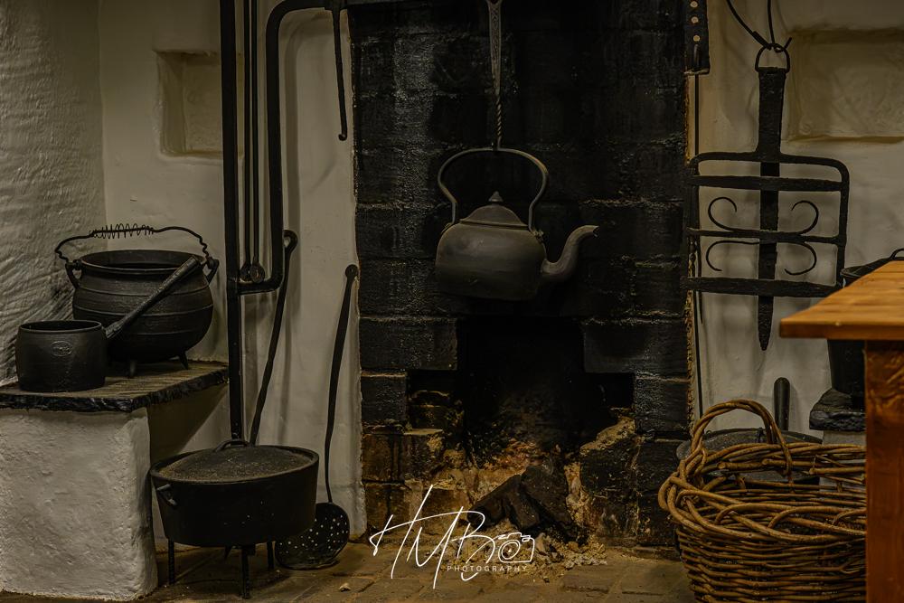 Some old black cast iron pots beside a smouldering peat fire. There is a kettle suspended on a crane hook over the fire. Other cooking and fire attending implements are visible. There is a woven basket to keep the turf in. Medieval heritage place.