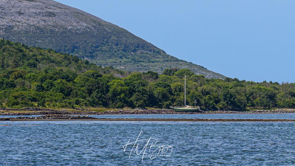 Another boat, parked up on some shingle, near Ballyvaughan Co Clare. There are plenty of trees and vegetation visible around it, a long bank of shingle reaches to the sea, and the sea is calm with small wavelets.