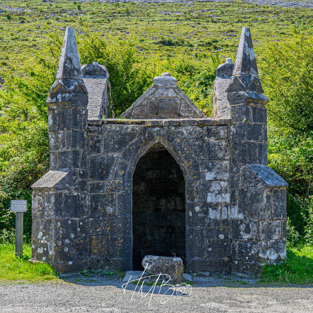 A small building protecting a communal water source. This was very important a few generations ago. It is a small gothic style construction, with four peaked towers, and an arched entrance. A green hill rises behind it.