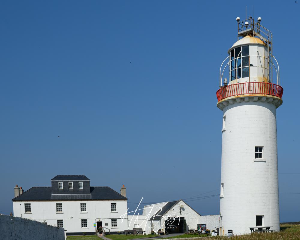 Hook Head lighthouse, with some of the accomodation buildings. Clear blue sky, a white tower with a red railing around the upper storey. The large magnifying glass to multiply the light can be seen in the upper story, facing to the left.
