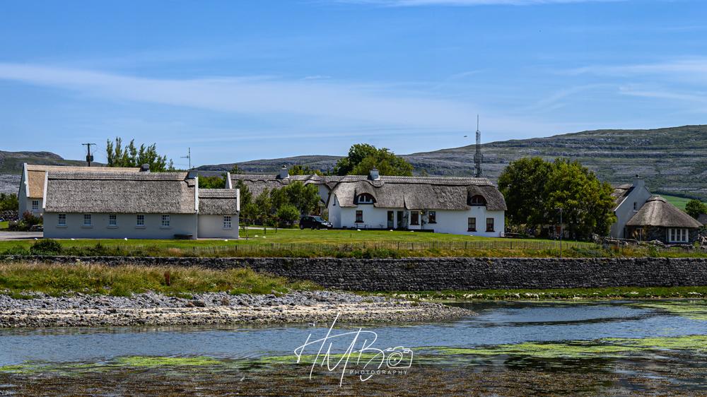 Rental cottages in a white render, with traditional thatched roofs, overlooking the bay at Ballyvaughan.  The stark stone of the Burren rises in layers behind to the right.