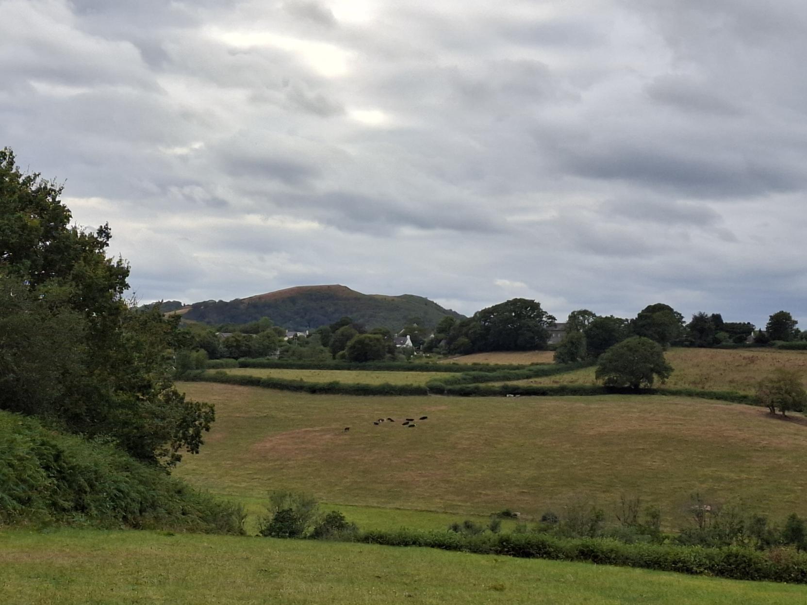 Across the fields and up the hill there is a hint of a village and above it a 2 peaked hill. The sky is cloudy but not rain filled