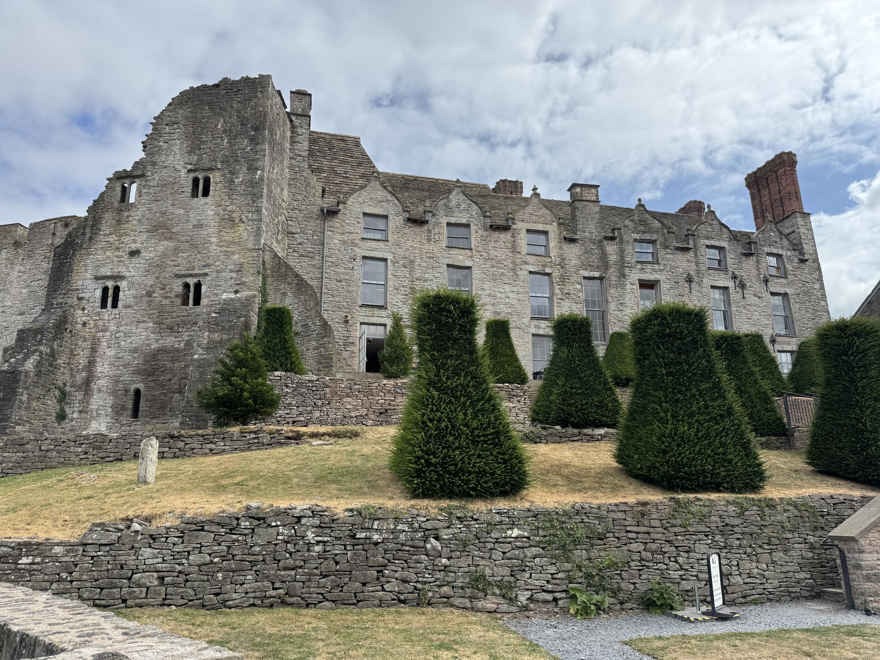 Hay on Wye castle.  A fortified Manor House built on a hill. The left facade is like a castle wall and partly ruined, whereas the right is an intact grey stone medieval manor.  There are large topiary hedges on the foreground and a grey stone wall.