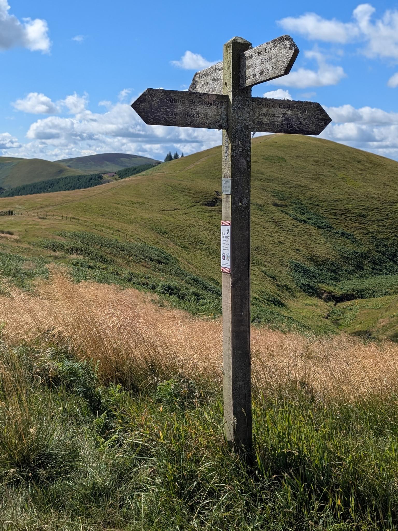 Colour photograph of a wooden finger post with four arrows pointing in different directions for different routes. It is standing on a stretch of grass in the background a hill rises away towards the distance. The sky is blue with fluffy white clouds.