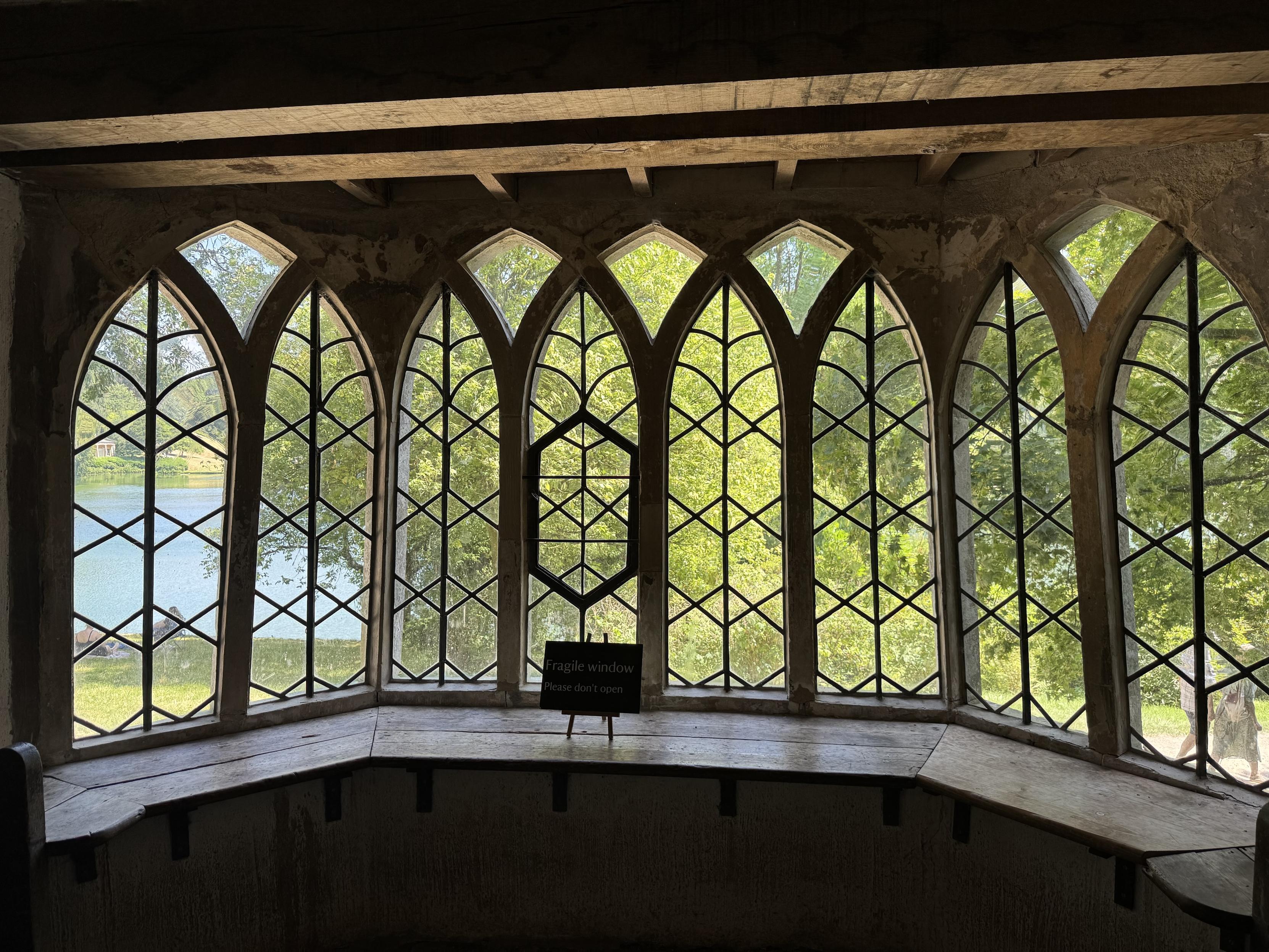 View from inside of bay window with Gothic arch windows in a 2-4-2 arrangement, with lead beading.  Through the windows you can just make out the lake and trees of the grand estate
