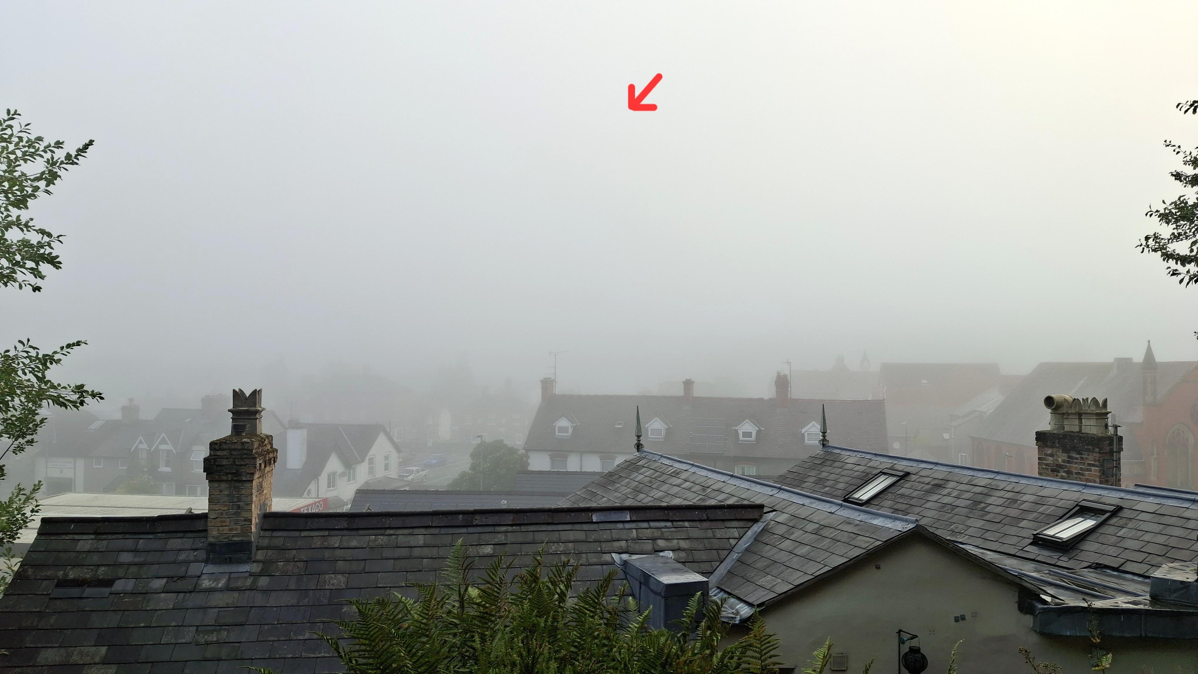 Damp rooftops of small Welsh town shrouded in heavy mist, with a tiny red arrow pointing to an invisible spot in the sky