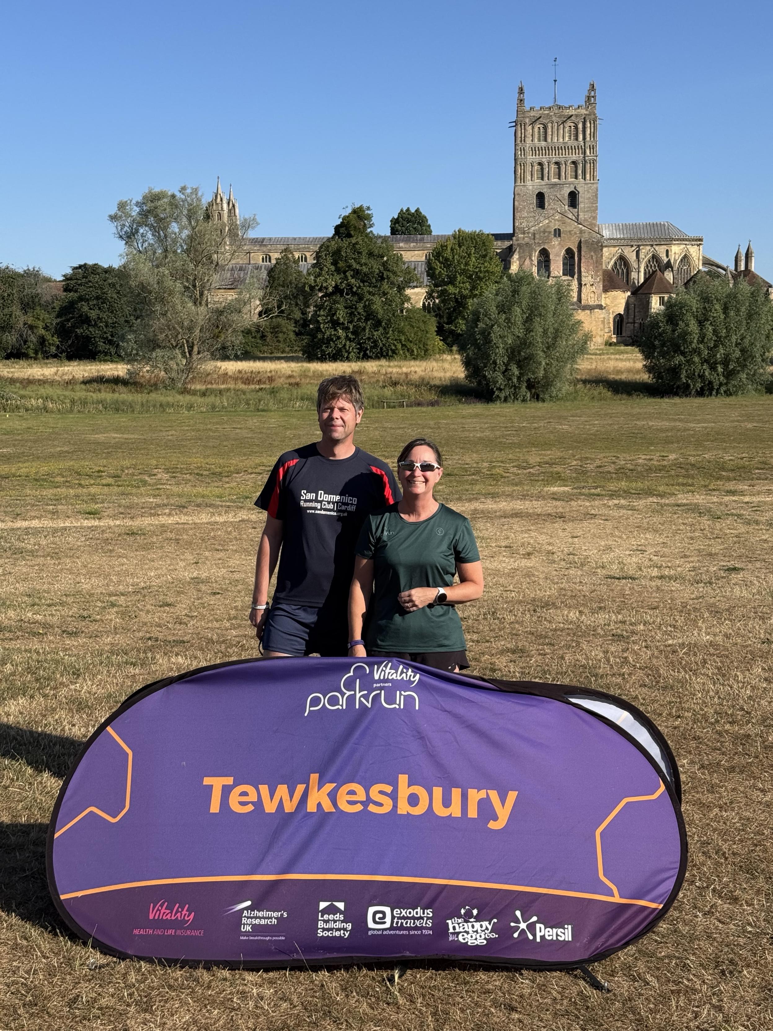 Ceri and I standing behind the Tewkesbury Parkrun sign in a field in front of Tewkesbury Abbey. The sky is pure blue and the sun is shining. The grass is very dry - golden in places.