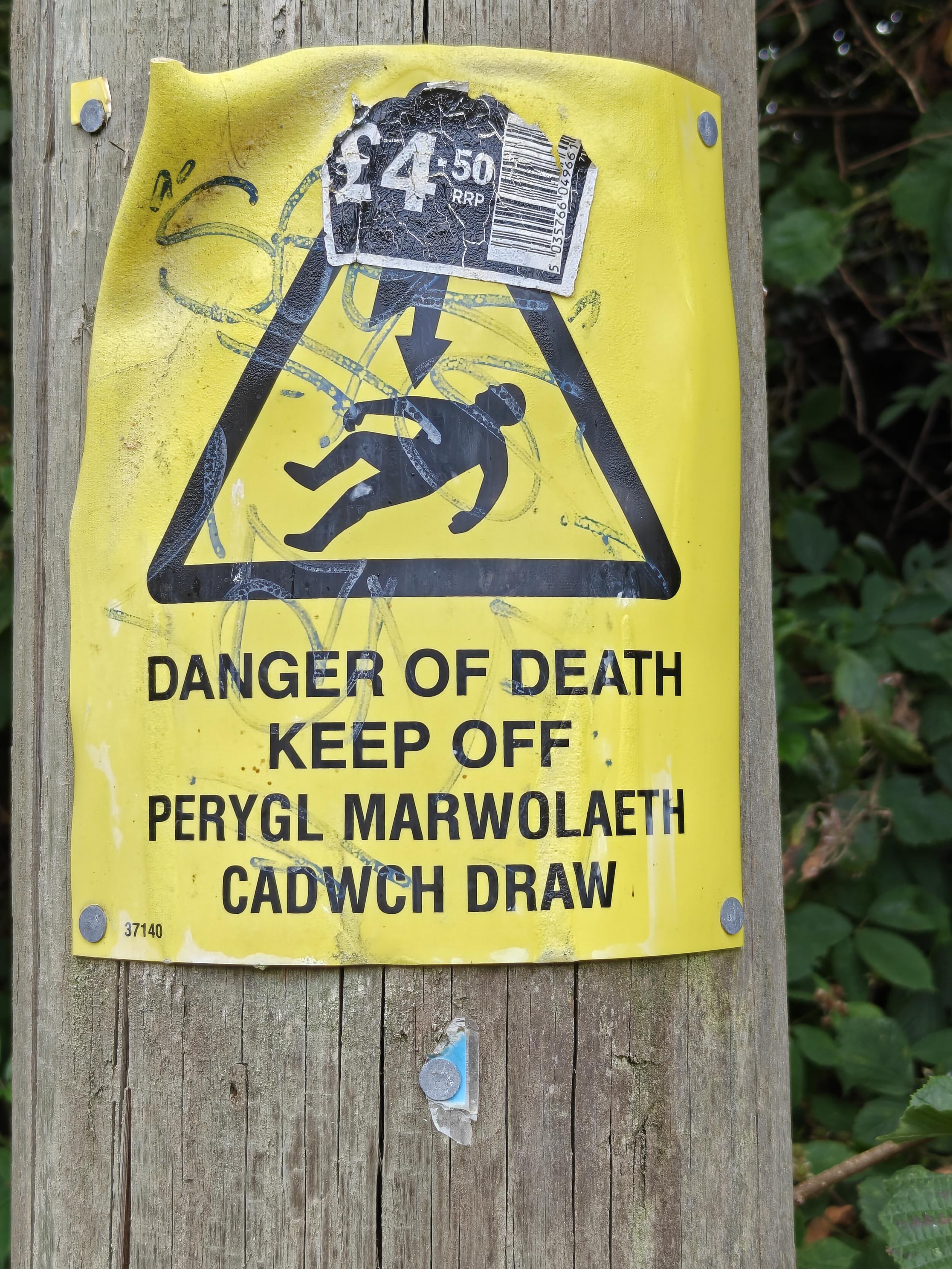 A hazard warning sign nailed to a pole carrying electric cables.  The sign is a black triangle on yellow background. Inside the triangle is an image of a body lying down above which is a zigzag arrow implying electric shock.
Underneath the triangle in block capitals is the bi-lingual (English/Welsh) message - DANGER OF DEATH KEEP OFF / PERYGL MARWOLAETH CADWCH DRAW.
The top of the triangle is obscured buy a supermarket price sticker that says £4.50 rrp.