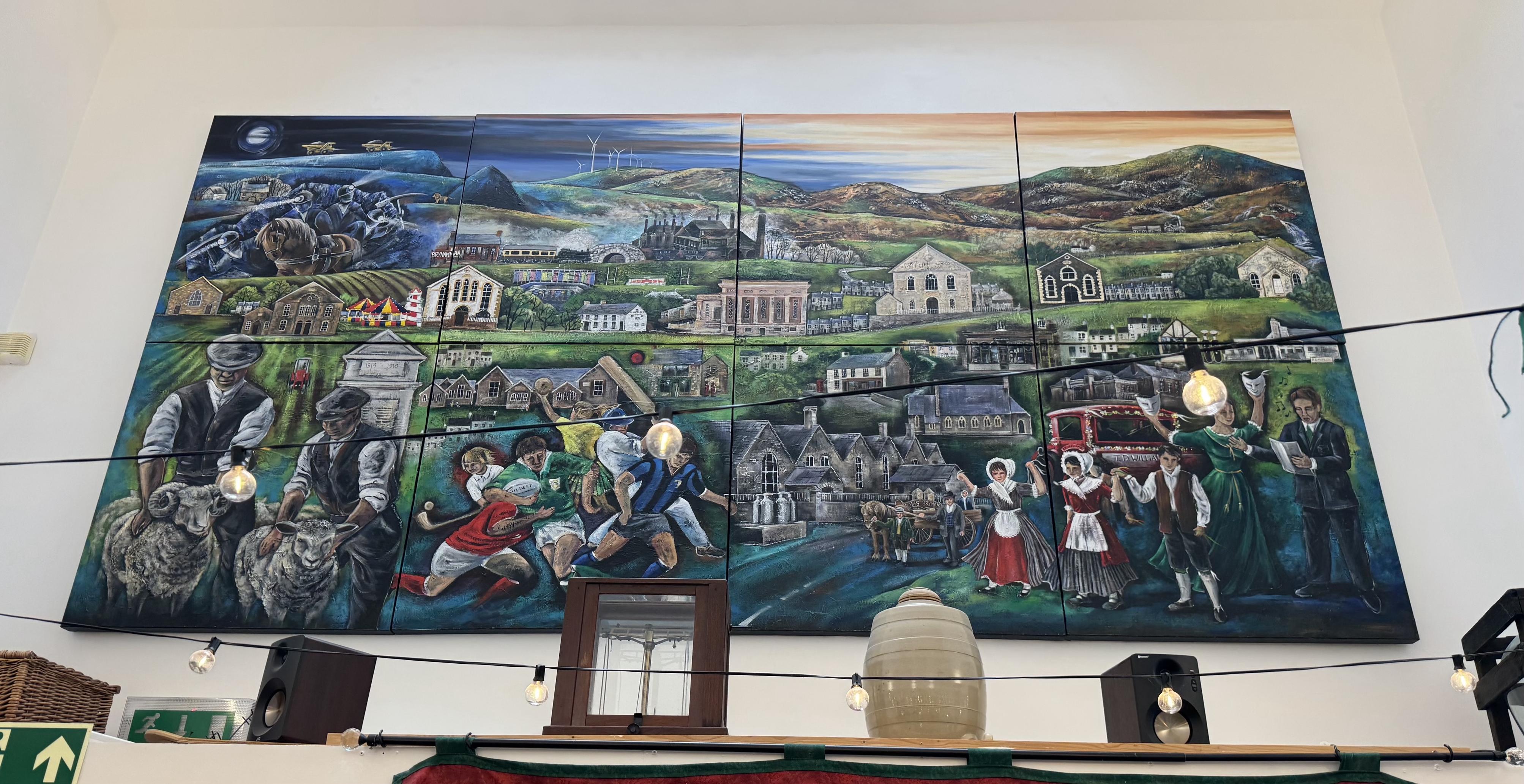 Large wall mounted mural of scenes from the local Welsh. Valley including villages,  boys playing sports, farmers with sheep, people in traditional Welsh costumes, chapels, terrace houses, rolling mountains and wind turbines 